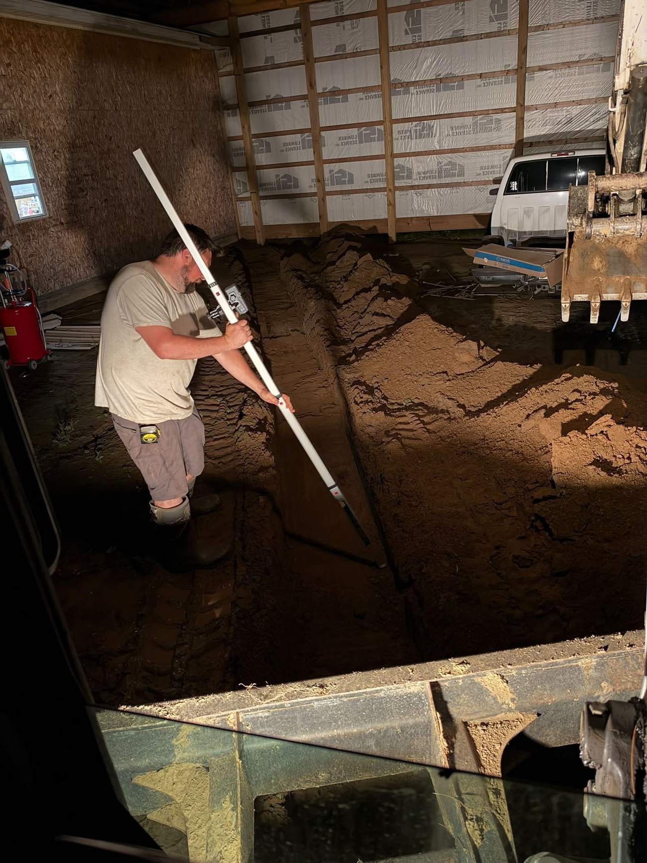 Grading and drainage work being laid out inside a garage foundation with an excavator staged outside